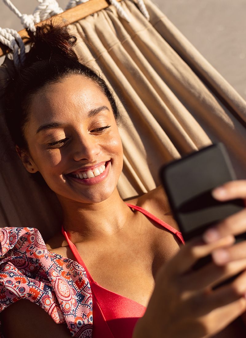 Vista frontal de una mujer usando un teléfono móvil mientras se relaja en una hamaca en la playa.